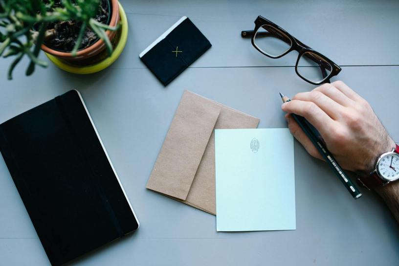 Overhead view of a desk with writing tools, glasses, envelope, and notebooks, showcasing office work essentials.