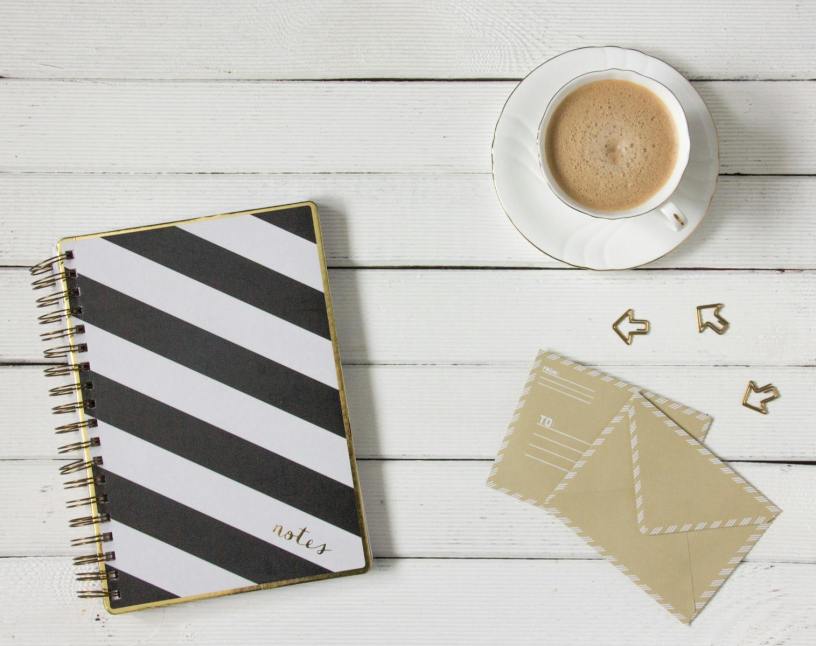 Flatlay of a striped notebook, coffee cup, and envelopes on a wooden desk.