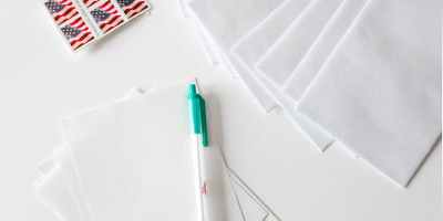 Flat lay of envelopes, blank paper, pen, and American flag stamps on white background.